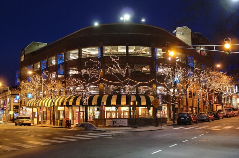 Decorated with string lights, a modern building stands at a street corner at night, showcasing its bustling atmosphere and inviting storefronts.