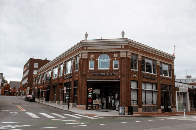 Historic brick building featuring "Wholesome Fresh" storefront, situated at a street corner with an overcast sky. Ideal for local shopping.